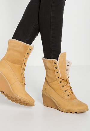 Bottes d'hiver beige à talon compensé avec doublure en fourrure et laçage à l'avant, portées avec un jean skinny noir sur un pied levé.