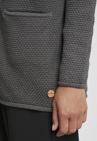 Gray textured sweater featuring a large pocket, wooden logo accent, and ribbed cuffs, paired with black fabric.