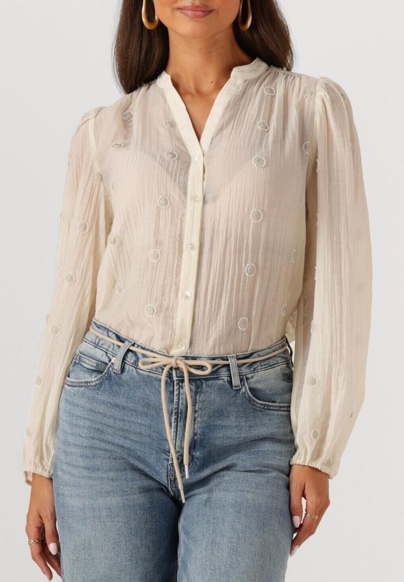 Vrouw in een doorschijnende crèmekleurige blouse met lange pofmouwen en parelachtige cirkel details, ingestopt in hoge taille blauwe jeans met een gestrikt stoffen riem.