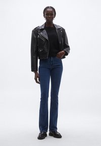 Jachetă de piele neagră de motociclist cu fermoare argintii, pulover negru și blugi evazați albaștri, asortați cu mocasini negri. Design simplu și bine mulat.