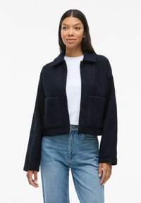 Marinblå kort jacka i texturerat tyg med krage, dragkedjestängning och två framfickor, bärs över en vit t-shirt och blå jeans.