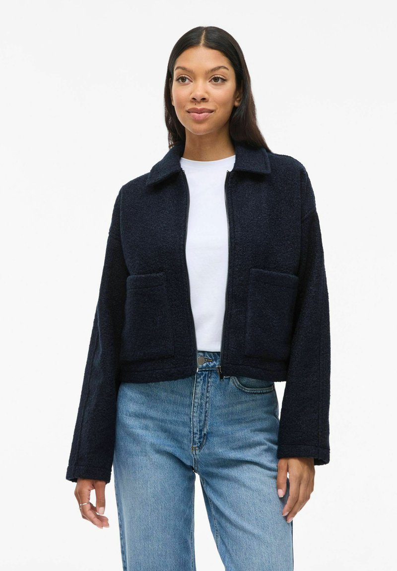 Marinblå kort jacka i texturerat tyg med krage, dragkedjestängning och två framfickor, bärs över en vit t-shirt och blå jeans.
