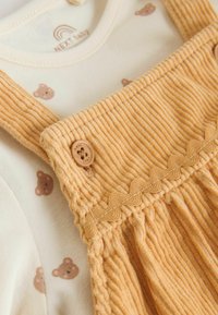 Gelber Cordoverall mit Rüschendetails und Knopfakzenten, über einem cremefarbenen Langarmshirt mit hellbraunen Bärenmustern getragen.