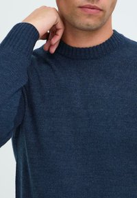 Nærbilde av en person som har på seg en mørkeblå strikket genser med ribbestrikket krage og mansjetter, hånden hvilende nær halsen.