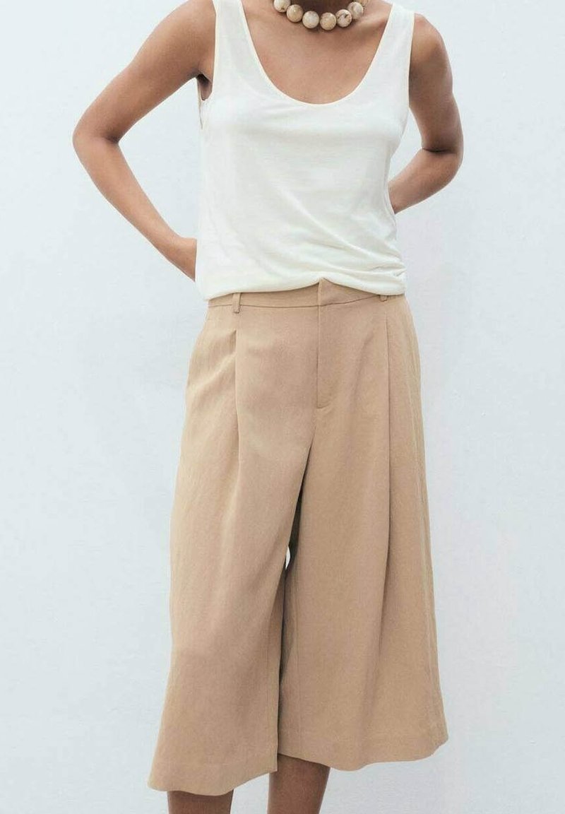 Pantalon beige à jambes larges avec des plis et une texture lisse, associé à un débardeur blanc à encolure ronde, et un collier de perles en bois.
