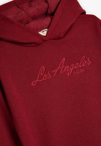 Felpa con cappuccio bordeaux realizzata in tessuto morbido, con logo ricamato "Los Angeles" in tonalità coordinata. Include un cappuccio regolabile con coulisse.