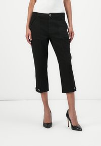Pantalones cortos negros de algodón con bolsillos frontales, cierre de botón y puños enrollados con botones a presión. Llevados con tacones negros.