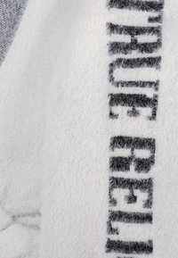 Weißer, flauschiger Pullover mit einer weichen Textur, der ein auffälliges Design mit schwarzen Buchstaben aufweist. Die Buchstaben erscheinen fett und leicht verschwommen.