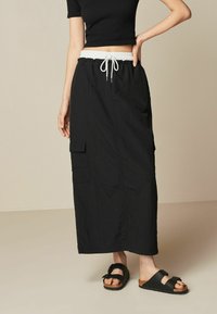 Zwarte maxi skirt met een elastische tailleband en trekkoord. Inclusief twee zijzakken. Draagt met zwarte sandalen met een kurken zool.