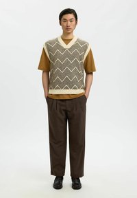Gilet tricoté à motif zigzag en marron et crème sur un t-shirt marron, associé à un pantalon ample marron et des chaussures noires.