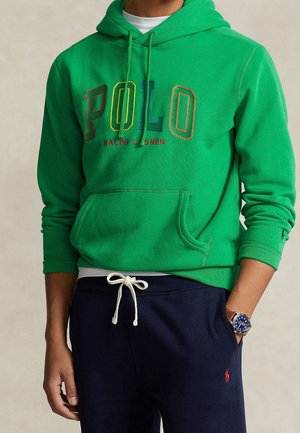 Osoba ubrana w zieloną bluzę z kapturem Polo Ralph Lauren z kolorowymi literami oraz granatowe spodnie ze sznurkiem i czerwonym logo, ręka w kieszeni, niebieski zegarek.