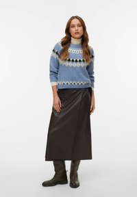 Ženska, oblečena v modri vzorčasti pulover čez bež puli majico, temno rjavo midi krilo in ujemajoče se rjave škornje, stoji pred enobarvnim ozadjem.