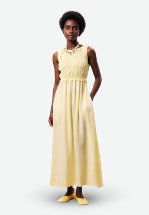 Femme aux cheveux courts et bouclés portant une robe maxi jaune sans manches et des chaussures plates jaunes assorties, debout avec une main dans la poche.