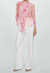 Blusa trasparente rosa con motivi floreali, con scollatura arricciata, abbinata a pantaloni bianchi a vita alta. Tessuto trasparente con una consistenza morbida.
