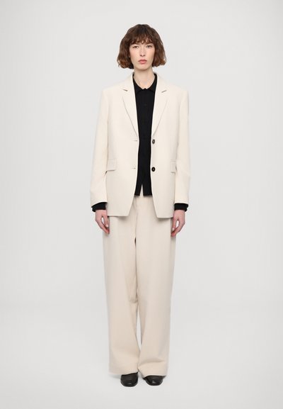 Beige übergroßer Blazer mit zwei Knöpfen und Revers, kombiniert mit weit geschnittenen Hosen. Schwarzes Hemd mit Knopfleiste darunter. Glatte Textur.