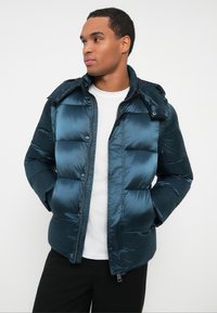 Hombre con una chaqueta puffer brillante en color verde azulado oscuro, con las manos en los bolsillos, camiseta blanca y pantalones negros, mirando hacia la izquierda sobre un fondo blanco.