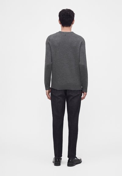 Homme debout, dos tourné, portant un pull gris à manches longues, un jean noir et des chaussures noires sur un fond blanc uni.