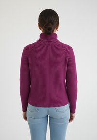 Fioletowy prążkowany sweter z golfem o długich rękawach i dopasowanym kroju, zestawiony z jasnoniebieskimi dżinsami. Materiał ma wyraźną fakturę.