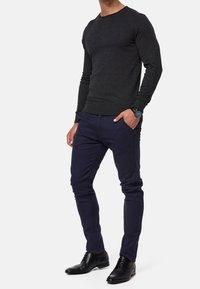 Mężczyzna w ciemnoszarym swetrze z długim rękawem, granatowych dopasowanych spodni, czarnych butach wizytowych i zegarku na ręku, stojący z jedną ręką w kieszeni.