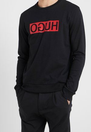 Personne portant un t-shirt noir à manches longues avec un logo rectangulaire rouge affichant le texte inversé « HUGO » sur la poitrine, mains dans les poches, sur fond uni.