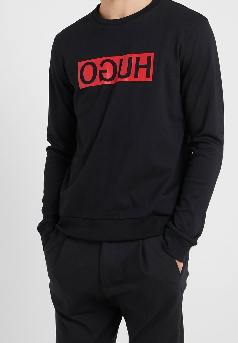 Personne portant un t-shirt noir à manches longues avec un logo rectangulaire rouge affichant le texte inversé « HUGO » sur la poitrine, mains dans les poches, sur fond uni.