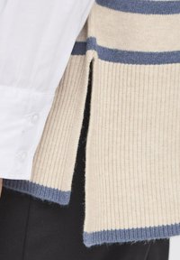 Beżowy sweter prążkowany w niebieskie paski, z bocznymi rozcięciami i miękką strukturą. Nosi się go na białej koszuli z detalem w postaci guzików.