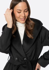 Černý, dvouřadý kabát s širokým límcem, texturovaný vlněný materiál, pásek v pase a knoflíkové detaily. Stylizovaný přes bílý rolák.