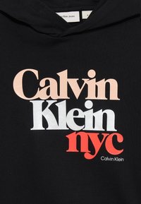 Čierna mikina s kapucňou s logom "Calvin Klein" v ružovej, bielej a červenej farbe. Obsahuje klokanie vrecko a kapucňu so sťahovacou šnúrkou. Mäkký bavlnený materiál.