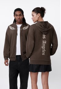 Smeđa zip-up dukserica s grafikom krila sprijeda i natpisom "Los Angeles" na leđima. Ima prostranu kapuljaču, kengurske džepove i opušteni kroj.