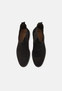 Pantofi din suede negri cu vârf rotund și panouri elastice pe lateral. Prezintă detalii brogue pe vârf și un mâner la călcâi.