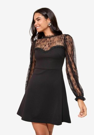 Kobieta uśmiechająca się, ubrana w czarną sukienkę z przezroczystymi koronkowymi długimi rękawami i koronkowym karczkiem nad dopasowanym gorsetem i spódnicą o kroju literą A.