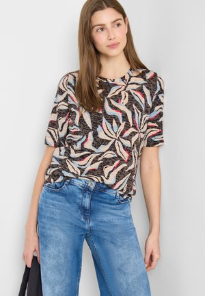 Jeune femme aux cheveux bruns lisses portant un haut à manches courtes à imprimé abstrait de feuilles et un jean bleu, tenant une veste noire dans une main.