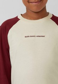 Crème en bordeaux raglan-sleeve sweatshirt met geborduurde tekst "SLVR STUDIOS | DEPARTMENT" in donkerbruin. Zachte katoenstof.