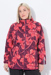 Žena s blond vlasy, která má na sobě vínový kabát s velkými červenými květinovými vzory, stojící proti bílé zdi.