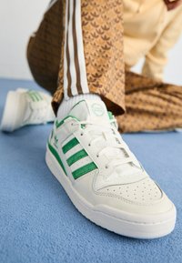 White sneaker with green stripes, textured leather, perforated toe box, and flat laces, set on a blue textured surface.