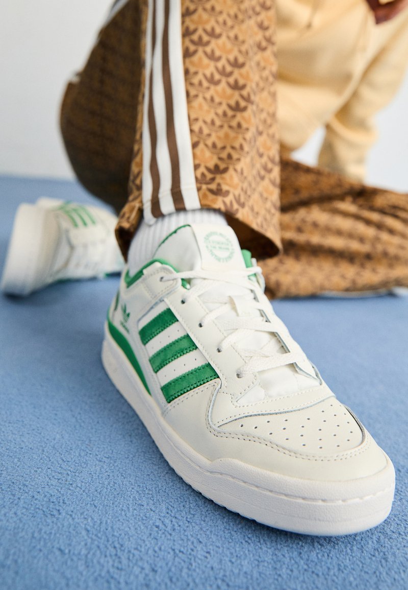 Zapatilla blanca con rayas verdes, cuero texturizado, puntera perforada y cordones planos, sobre una superficie azul texturizada.