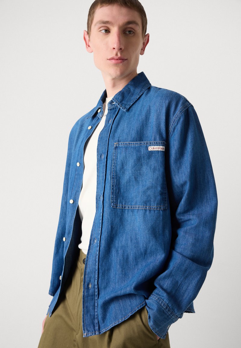 Denimhemd, blau, mit Knöpfen und Kragen, einer Brusttasche, weißen Nähten und Logopatch. Kombiniert mit beigefarbenen Hosen.