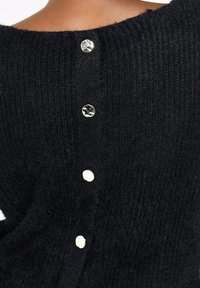 Černý, chlupatý svetr s širokým výstřihem a pěti lesklými, kulatými stříbrnými knoflíky na zadní straně. Texturovaná pletenina.