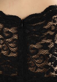 Tissu en dentelle noire avec motif floral, doté d'une fermeture éclair discrète à l'arrière. La texture est délicate et légèrement transparente.