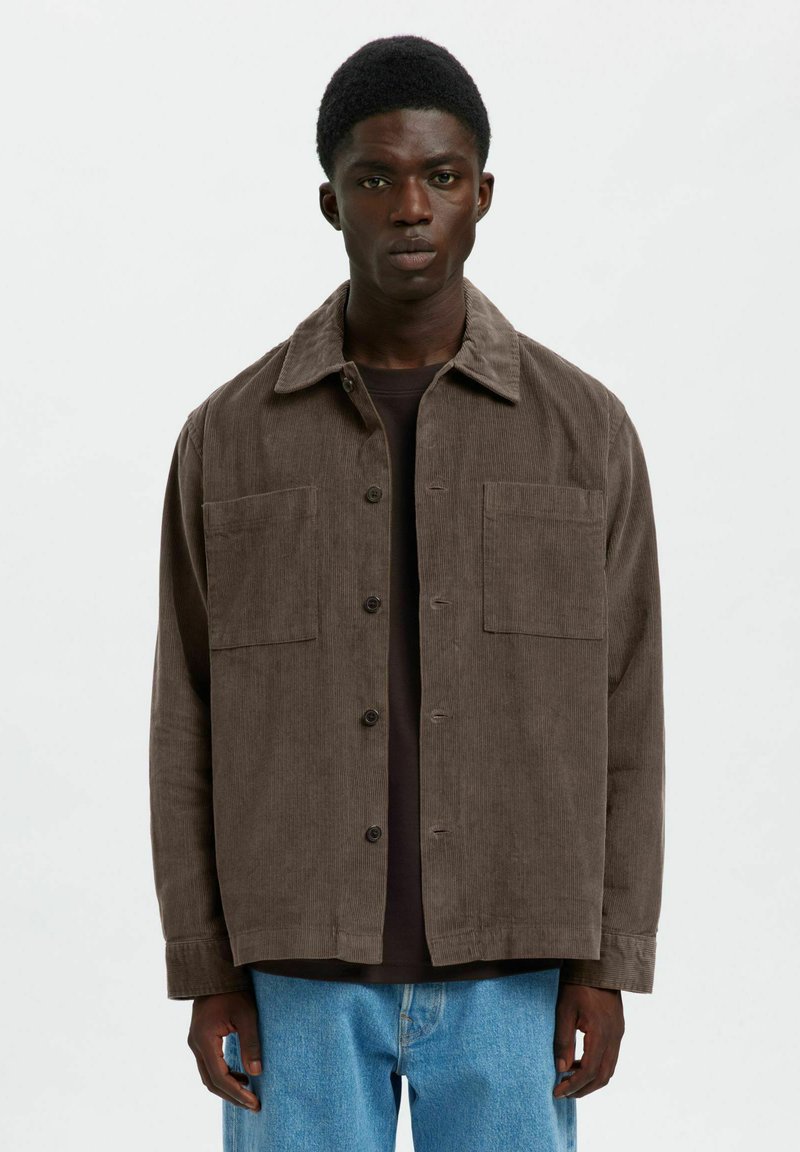 Hombre de piel oscura que lleva una chaqueta de pana marrón holgada sobre una camisa negra y jeans azul claro, de pie contra un fondo blanco plano.