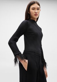 Žena oblečená v priliehavom čiernom rebrovanom svetri s perovými manžetami a čiernych nohaviciach s vysokým pásom, stojaca s jednou rukou v vrecku.