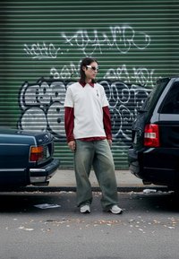 White t-shirt layered over red long-sleeve shirt, loose light blue jeans, and white sneakers. Sleek sunglasses, urban graffiti background.