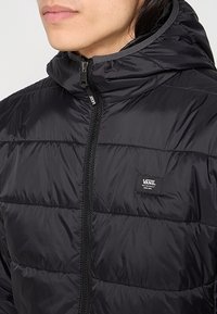 Czarna kurtka puchowa z wysokim kołnierzem, zamkiem błyskawicznym i poziomymi przeszyciami. Widoczna charakterystyczna naszywka z logo Vans na piersi.