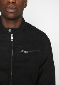 Zwarte suède jas met een hoge kraag, een ritssluiting aan de voorkant en een klein ritszakje op de borst met metalen accenten.