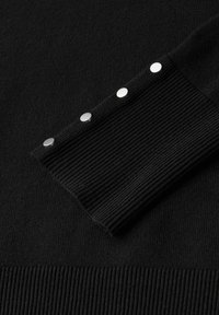 Polsino di un maglione nero lavorato a maglia con texture a coste e tre bottoni a pressione argentati per la chiusura su un materiale liscio.