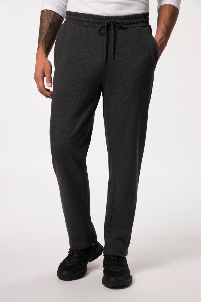 Uomo con pantaloni della tuta grigio scuro con coulisse e sneakers nere, in piedi con le mani nelle tasche davanti a uno sfondo chiaro e uniforme.