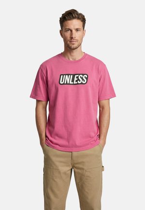 Uomo che indossa una T-shirt rosa con la scritta "UNLESS" e pantaloni cargo beige, in piedi con le mani nelle tasche su sfondo bianco.