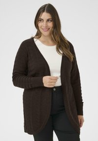 Dunkelbrauner gestrickter Cardigan mit Zickzack-Muster, offener Front und gerippten Kanten. Getragen über einem weißen Oberteil, kombiniert mit schwarzen Hosen.
