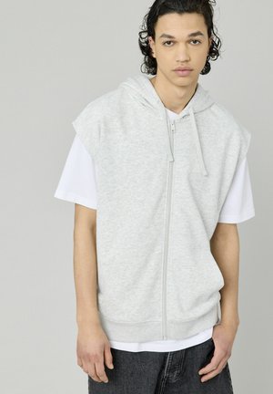 Haut sans manches gris clair avec une fermeture éclair à l'avant, une capuche à cordon de serrage et des poches kangourou, porté par-dessus une chemise blanche à manches courtes et un jean noir.