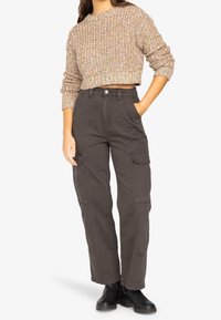 Pantalon cargo marron avec poches latérales et jambes droites, associé à un pull court en maille texturée beige. Bottines noires complètent la tenue.